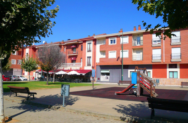 Parkszene mit Spielplatz, Bänken, Bäumen, Gras und umgebenen Gebäuden, mit einer Gruppe von Menschen unter einem Dach, Zelten, Fahrzeugen und einem bewölkten Himmel.