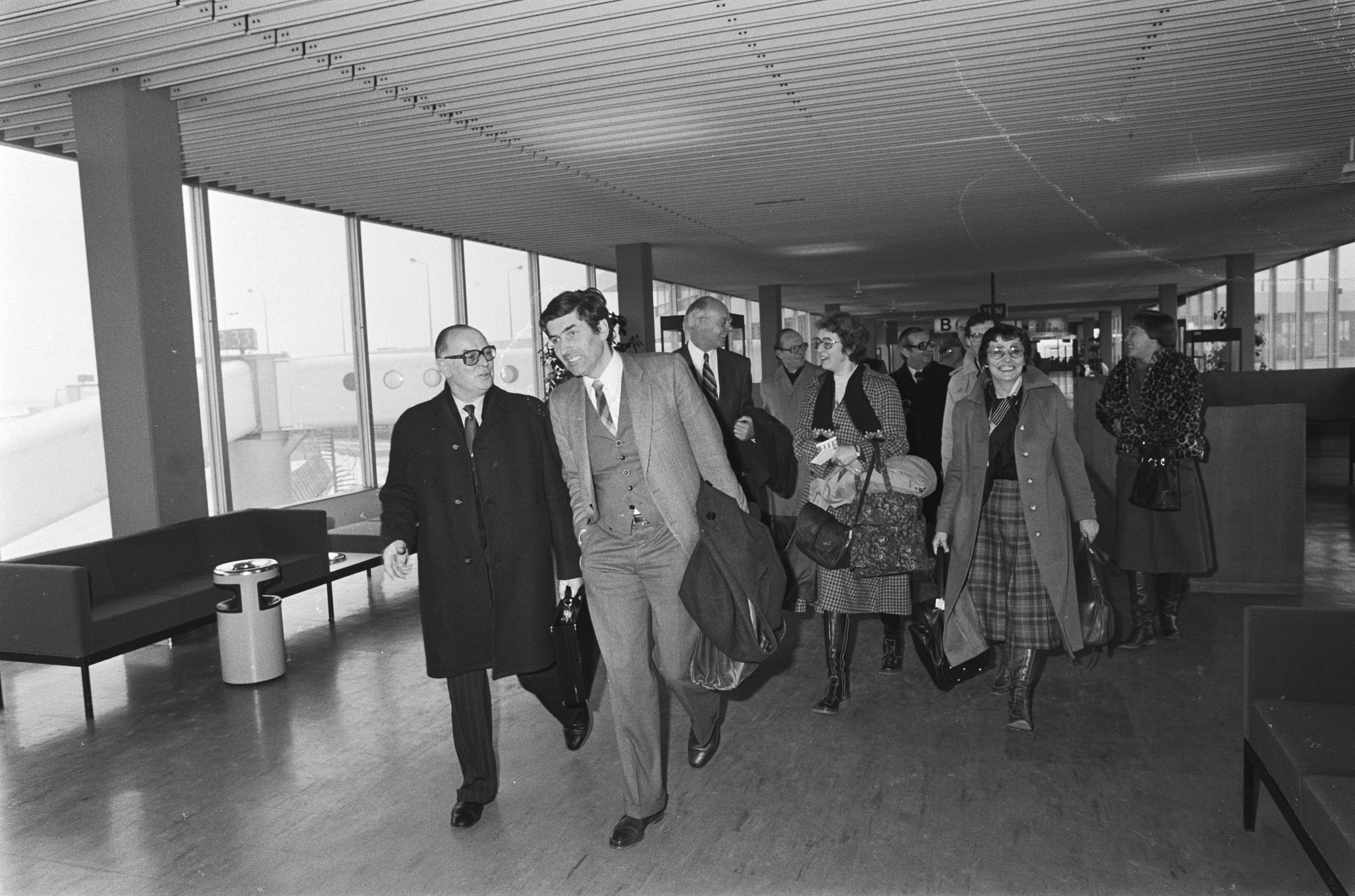Schwarz-weißes Bild von Menschen, die durch einen Flughafen gehen und Gepäckstücke, Sofas, einen Müllcontainer, Säulen und Glasfenster im Hintergrund tragen.