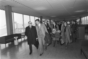 Schwarz-weißes Bild von Menschen, die durch einen Flughafen gehen und Gepäckstücke, Sofas, einen Müllcontainer, Säulen und Glasfenster im Hintergrund tragen.