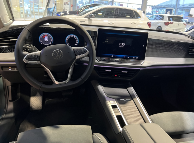 Innenansicht eines Volkswagen e-Golf mit Lenkrad, Armaturenbrett, Bildschirmen, Knöpfen, Sitzen und Glastüren, mit Blick auf andere Autos, Gebäude, Bäume, Pfähle und den Himmel draußen.