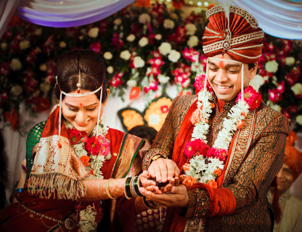 A man and woman in traditional Indian wedding attire exchange rings as guests and floral decorations are visible in the background under a curtain.