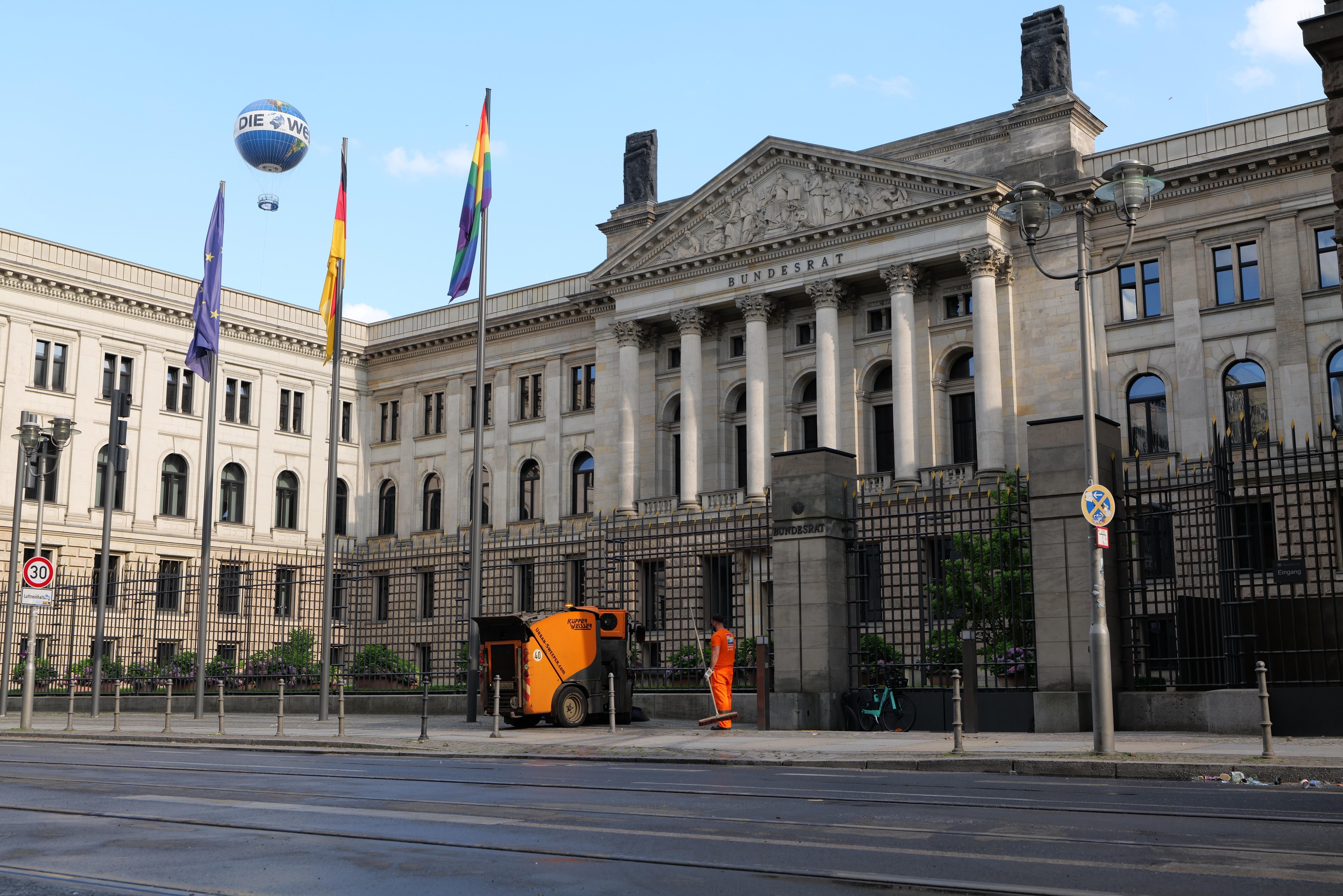 Large modern building with many windows, identified as the Bundestag in Berlin, surrounded by street infrastructure, pedestrians, flags, and trees under a cloudy sky.