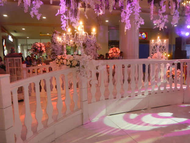 Eine Hochzeitsfeier mit einer weißen Balustrade, die mit Blumen und von der Decke hängenden Kronleuchtern geschmückt ist, viele Menschen drum herum, Säulen und Lichter im Hintergrund und ein Bildschirm auf der rechten Seite.