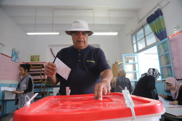 Ein Mann mit Hut stimmt an einer Wahllokal, vor einer roten Wahlurne mit einem Stück Papier in der Hand stehend, ab, während andere an Tischen sitzen und schreiben, mit einem Fenster und einem Vorhang auf der rechten Seite.