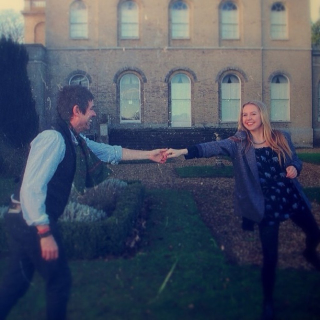 Ein Paar steht auf einer grünen Fläche, hält sich an den Händen, mit einem Gebäude und Bäumen im Hintergrund.