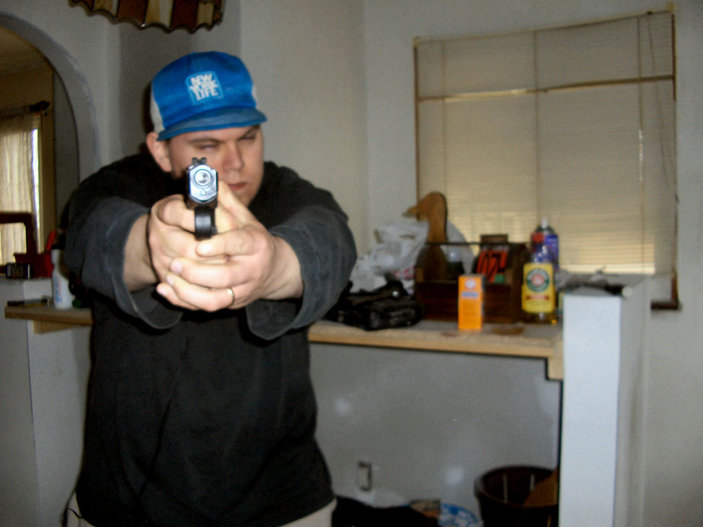 Ein Mann hält eine Pistole mit beiden Händen, steht vor einem Tisch mit verschiedenen Gegenständen, mit einer Wand und zwei Fenstern im Hintergrund.