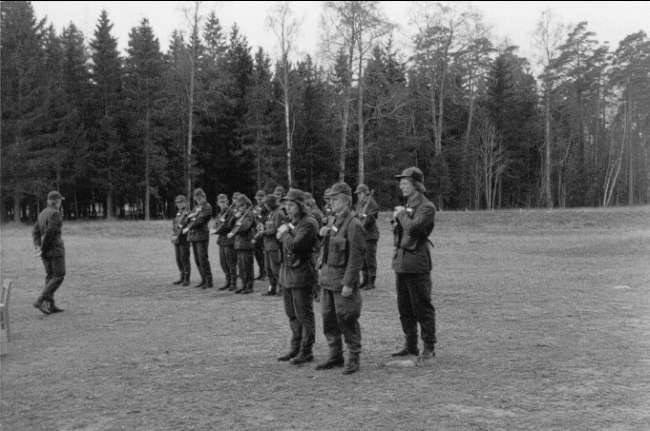 Schwarzes und weißes Bild einer Gruppe von Männern in militärischer Kleidung, die in einem Feld stehen und Gewehre halten, mit Bäumen und einem klaren Himmel im Hintergrund, wahrscheinlich während einer Übung.