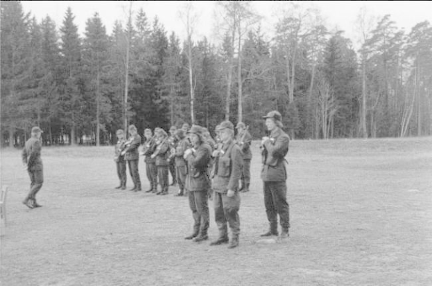 Schwarzes und weißes Bild einer Gruppe von Männern in militärischer Kleidung, die in einem Feld stehen und Gewehre halten, mit Bäumen und einem klaren Himmel im Hintergrund, wahrscheinlich während einer Übung.