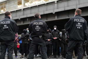 Eine Gruppe uniformierter Polizisten steht vor einer Menge von Menschen in schwarzen Uniformen und Masken, mit einer Brücke und einem Gebäude im Hintergrund, während einer Protestaktion in einer Stadt.