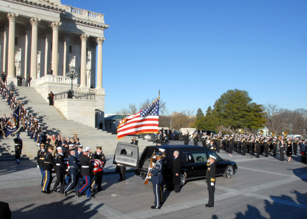 Funeral procession in front of the U.S. Capitol Building with people in caps holding flags, vehicles, steps with railings, light poles, trees, and sky visible.