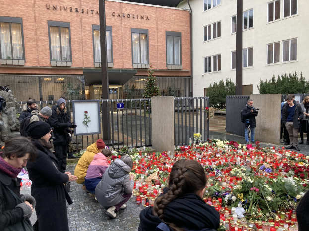 A group of people gathers around flowers and candles placed on the ground outside a building, some wearing caps or holding cameras and bouquets, with buildings, trees, and a board with text in the background.