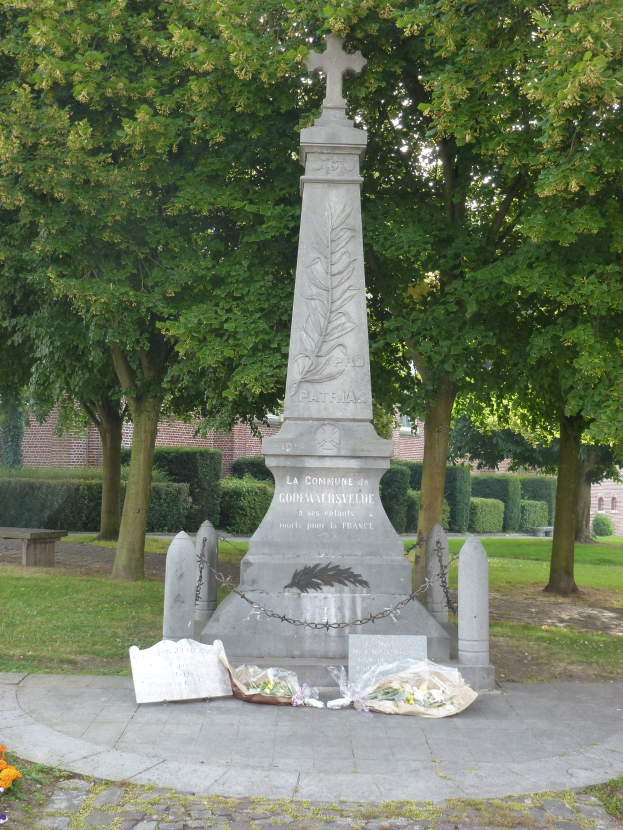 Kriegsdenkmal in einem Park umgeben von Bäumen und Pflanzen, mit einem Gebäude im Hintergrund und Blumen am Boden als Erinnerung.