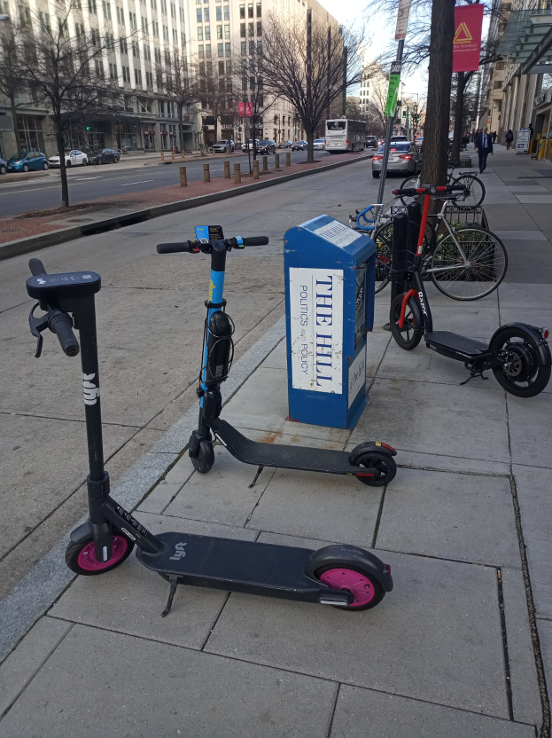 Zwei Elektro-Scooter auf einer städtischen Straße geparkt mit Fahrrädern, einem Müllcontainer und anderen städtischen Elementen im Vordergrund und Fahrzeugen, Bäumen, Gebäuden und Menschen im Hintergrund.