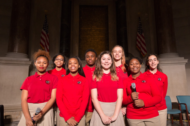 Eine Gruppe junger Frauen in roten Shirts, die strahlend Mikrofone halten und vor Stühlen, Säulen, Flaggen und einer Wand stehen, wahrscheinlich Teil eines nationalen Jugendchors.