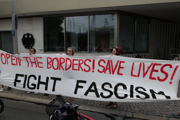 Group of people holding a banner reading "Open the Borders, Save Lives, Fight Fascism" in front of a building with glass windows and a pole, with a bicycle and bag on the road.