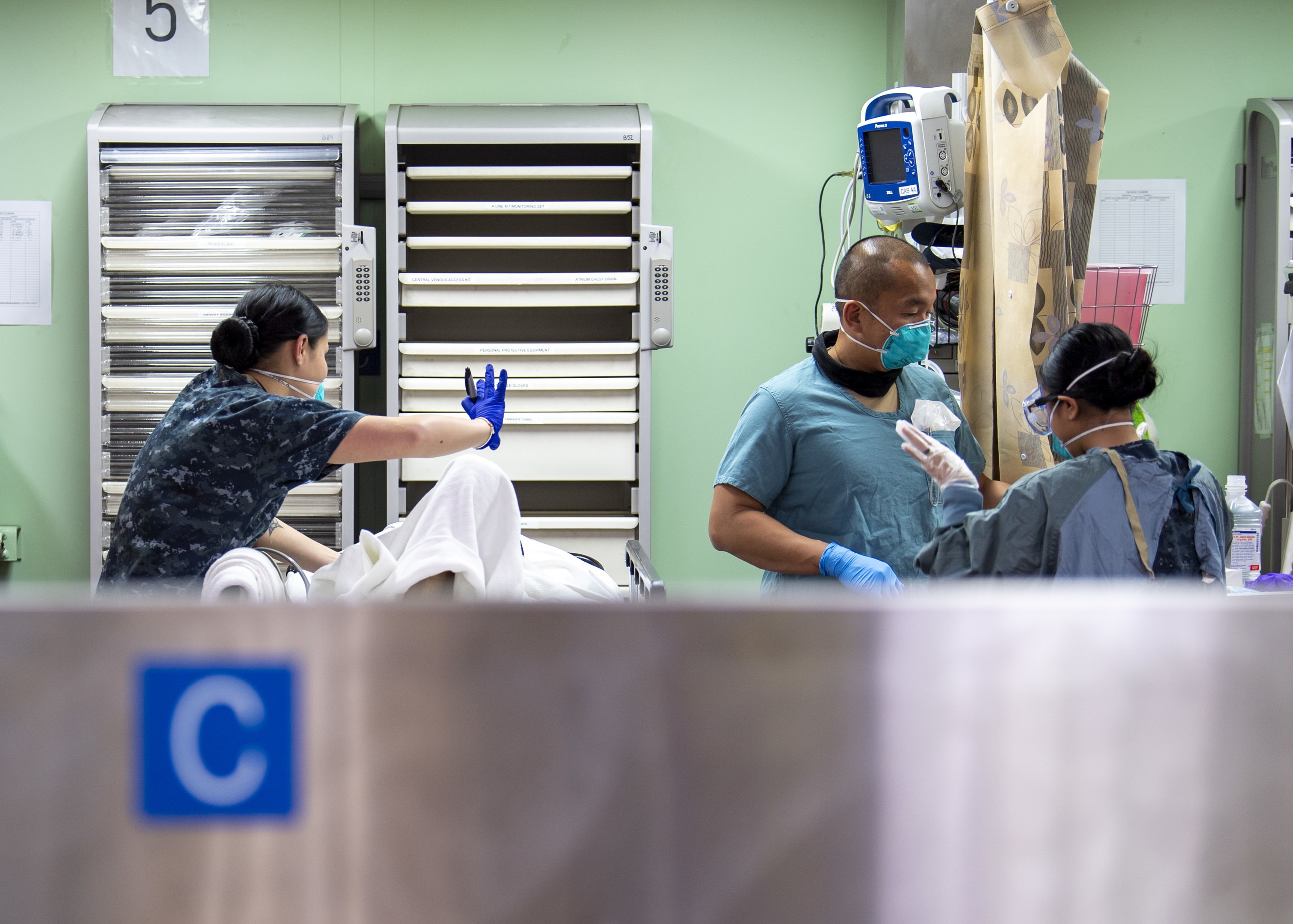Three healthcare workers in scrubs and masks attend to a patient lying on a hospital bed draped with a white cloth, with medical supplies on a nearby table and equipment visible in the background.