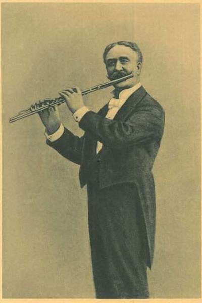 Old black-and-white photo of a man in a tuxedo playing a flute on stage, eyes closed in concentration, with slicked-back hair and stage lighting illuminating his face.