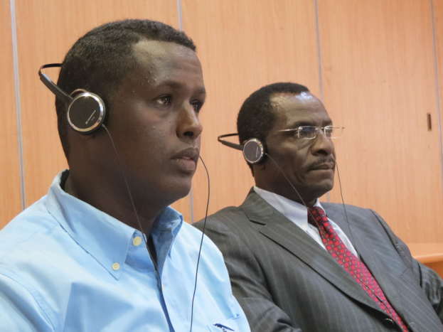 Zwei Männer in einem Gerichtssaal mit Kopfhörern, einer trägt ein blaues Hemd und der andere eine Brille, vor einer hölzernen Wand sitzend.
