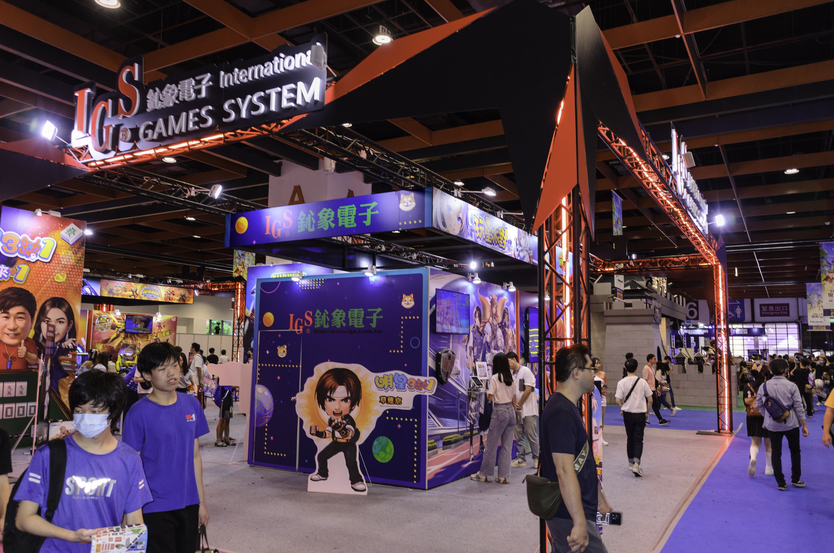 A group of people walking through a convention hall at a gaming expo in China, some carrying bags and holding objects, with text and image boards, ceiling lights, and carpeted flooring.