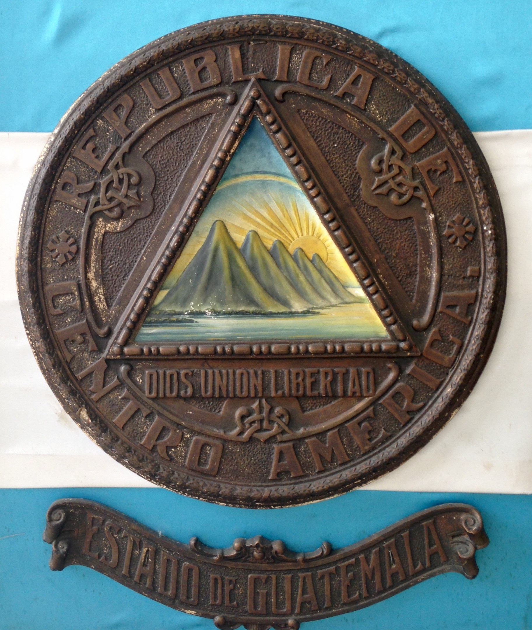 Plaque on a building with the Guatemalan flag in the background, featuring inscribed text.