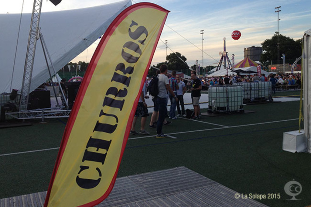 Lebendige Faschingsstimmung mit Zelten, zentralem Mast, Menschen, Bäumen, Strommasten, Straßenbahnen auf der Straße und einem Eisenständer auf der linken Seite unter einem klaren Himmel.