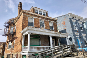 Eine Reihe verfallener Häuser an einer Straßenecke, mit zerbrochenen Fenstern, bröckelnden Wänden, Geländern, Stufen, einem Zaun, einer Schüsselantenne, Drähten und einem bewölkten Himmel im Hintergrund.