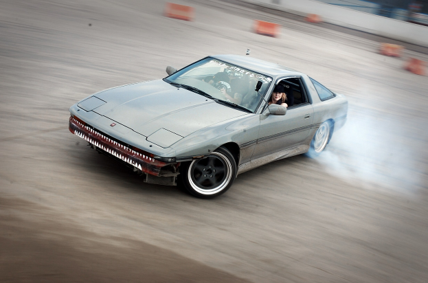 Eine Person in einem Helm fährt ein Auto, das Rauch auf einer von orangefarbenen Kegeln umgebenen Strecke ausstößt, mit einem unscharfen Hintergrund, der hohe Geschwindigkeit andeutet.