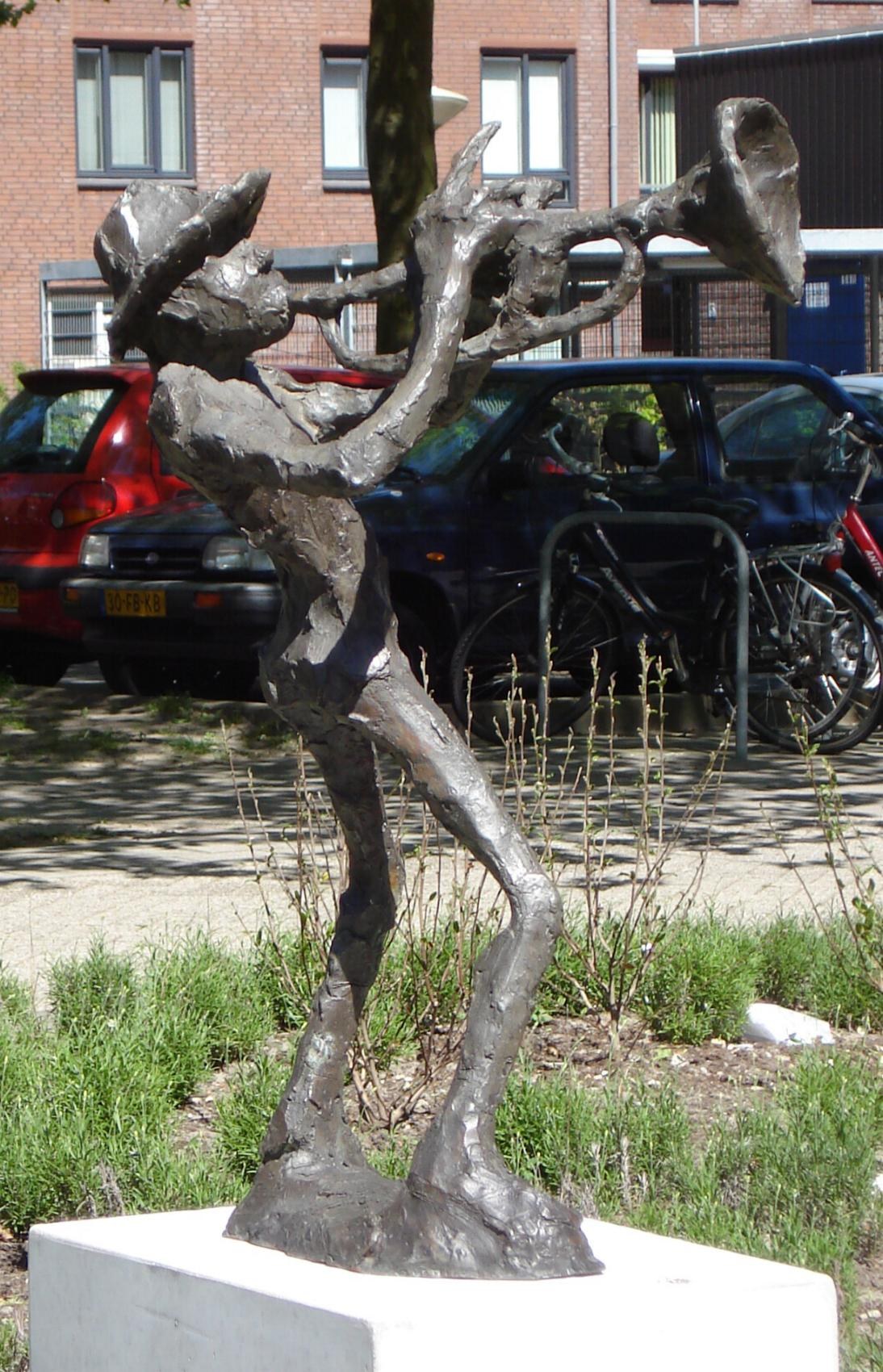 Bronzestatue eines Mannes, der eine Trompete spielt, auf einem weißen Sockel, umgeben von Gras und Pflanzen, mit Fahrzeugen, einem Baum und einem Gebäude im Hintergrund.