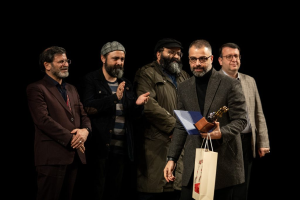 Eine Gruppe Männer steht auf einer Bühne, einer hält eine Tasche und eine Trophäe, mit dunklem Hintergrund, der die Eröffnungsnacht des Berliner Filmfestivals suggeriert.