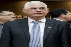 Ein Mann in einem blauen Anzug, weißem Hemd und gemustertem Schlips steht vor einer Gruppe von Menschen und spricht mit ernster Miene.