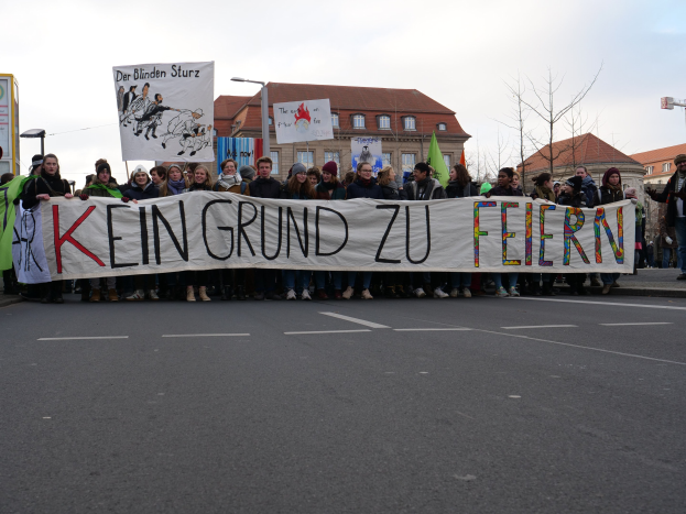 Gruppe von Menschen mit einem Transparent, das 'Kein Grund zu Feiern' trägt, bei einer Demonstration gegen Sparmaßnahmen, mit Gebäuden, Bäumen und einem klaren Himmel im Hintergrund.