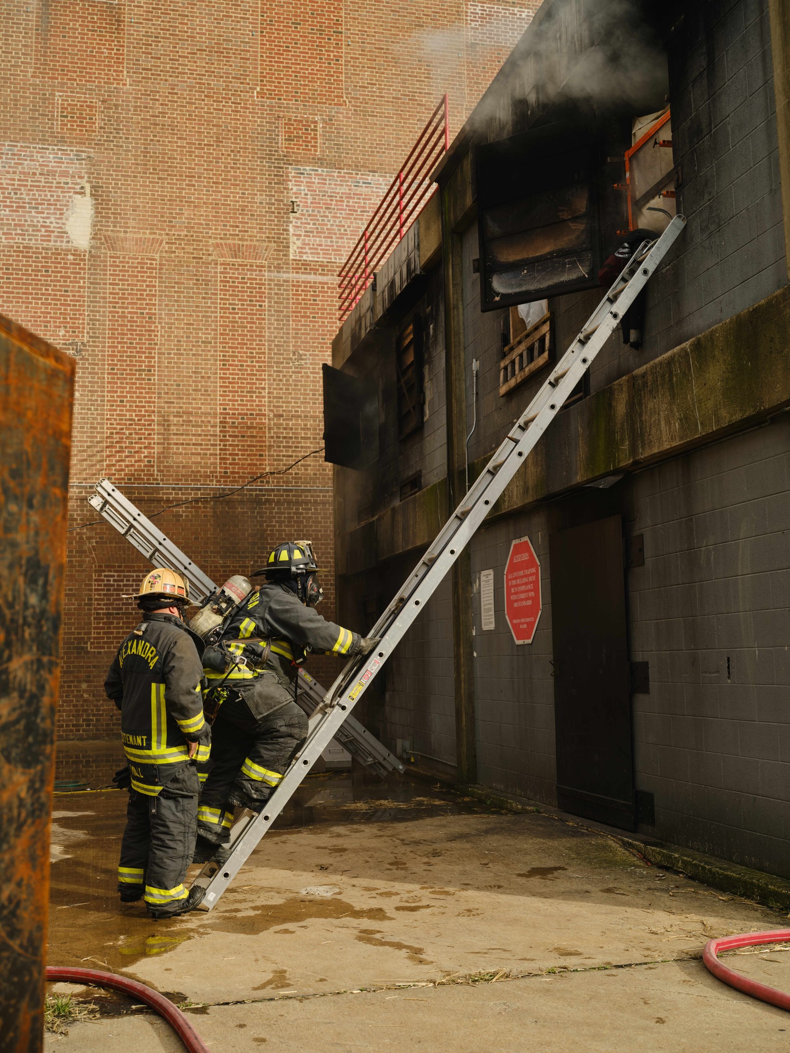 Feuerwehrleute in Helmen, die eine Leiter zu einem raucherfüllten Gebäude hinaufsteigen, mit einem metallischen Objekt links und Rohren am Boden.
