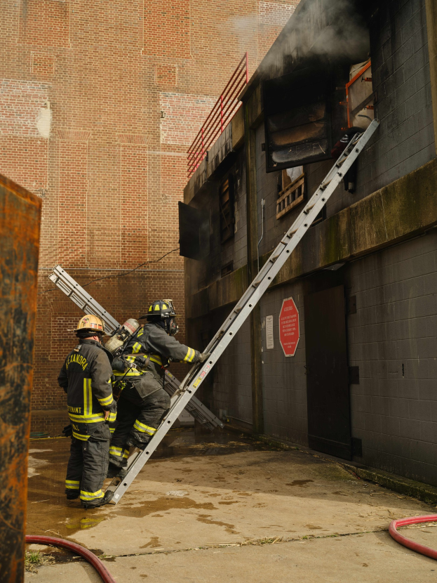 Feuerwehrleute in Helmen, die eine Leiter zu einem raucherfüllten Gebäude hinaufsteigen, mit einem metallischen Objekt links und Rohren am Boden.