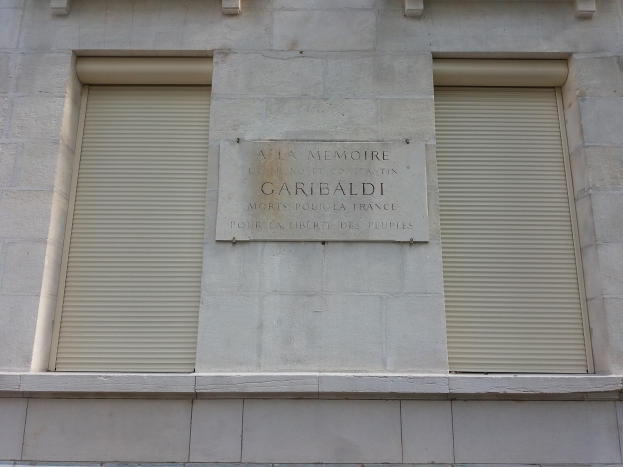 Ein Gebäude mit zwei Fenstern und Läden sowie einer Tafel an der Seite, wahrscheinlich ein Denkmal.