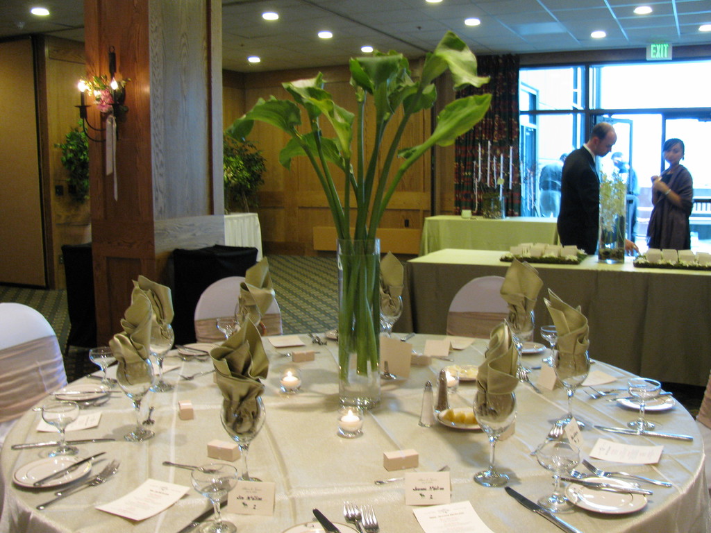 Ein Tisch mit einer weißen Decke, Tellern, Besteck, einem Glas und Baumzweigen, umgeben von Stühlen, mit einem grünen Tisch dahinter, einem Mann in einem schwarzen Blazer, einem stehenden Mädchen, einer Ausgangstür und zwei Männern hinter der Tür.