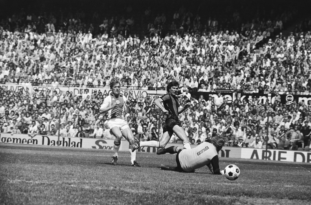 Schwarzes Foto von Männern, die Füßball auf einem Feld mit Zuschauern im Hintergrund und einem Banner mit der Aufschrift "Bundesliga 1978-79 Ajax 1-1 Hannover 96" spielen.