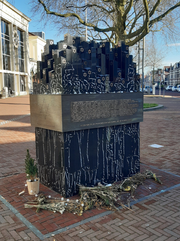 Denkmal für Holocaust-Opfer in einem Stadtplatz mit einem Baum davor, einem Topf mit einer Pflanze, verstreuten trockenen Blättern und Gebäuden im Hintergrund.