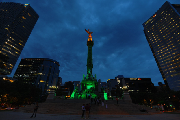 Gruppe von Menschen vor einem beleuchteten Denkmal in Mexiko-Stadt bei Nacht, mit Treppen, Bäumen und Gebäuden im Hintergrund.