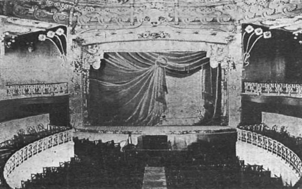 Schwarzes und weißes Foto des Theatersaals des Theatre Royal in London, das eine Bühne mit einem Vorhang, Geländern und ornamentalen Wanddesigns zeigt, mit einigen sitzenden Menschen und Text am unteren Rand.