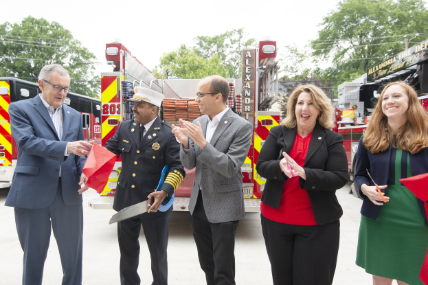 Eine Gruppe von Menschen steht vor einem Löschfahrzeug, klatscht und lacht, wobei zwei eine Schere und ein rotes Tuch halten, während eine Ribbon-Cutting-Zeremonie für die Alexandria Fire Department gefeiert wird, mit Bäumen, Drähten und einem klaren blauen Himmel im Hintergrund.