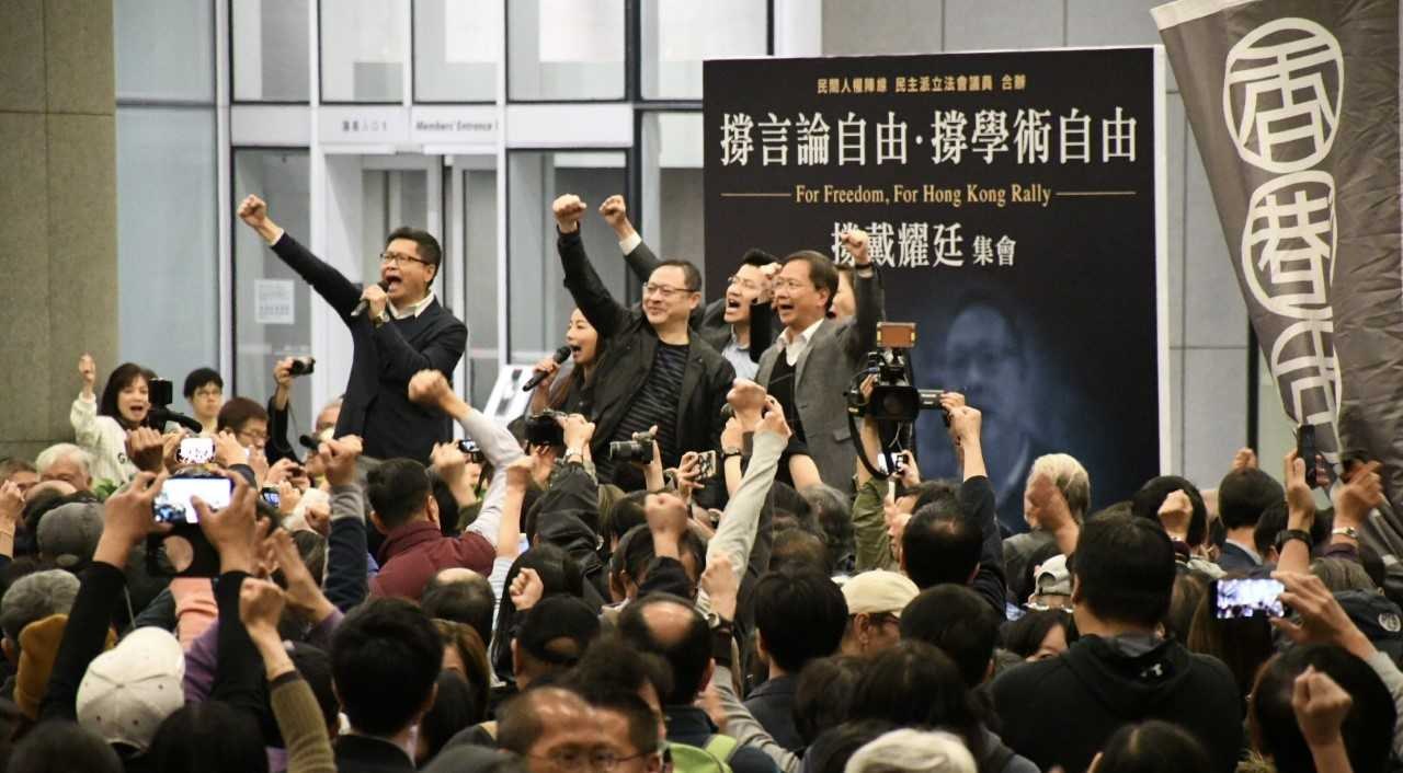 A group of people stands before a crowd holding cameras, phones, and microphones, with a banner, wall, and glass doors in the background, suggesting a protest against Hong Kong's extradition bill ban.
