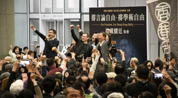 A group of people stands before a crowd holding cameras, phones, and microphones, with a banner, wall, and glass doors in the background, suggesting a protest against Hong Kong's extradition bill ban.