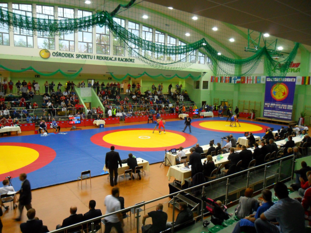 Großes Turnhalle mit Zuschauern bei einem Wrestling-Match, sitzende und stehende Zuschauer, Tische mit Gegenständen, Banner an den Wänden und beleuchtete Deckenverzierungen.