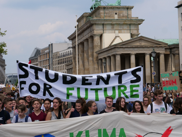 Eine Gruppe von Schülern marschiert in Berlin, eine bunt bemalte "Students for Future"-Fahne schwingend, vor einer Kulisse aus Gebäuden, Bäumen und Himmel.