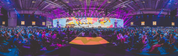 Eine große Zuschauermenge sitzt auf Stühlen einem hell erleuchteten Bühne mit Bildschirmen gegenüber, auf der mehrere Personen stehen während eines Online-Marketing-Events.