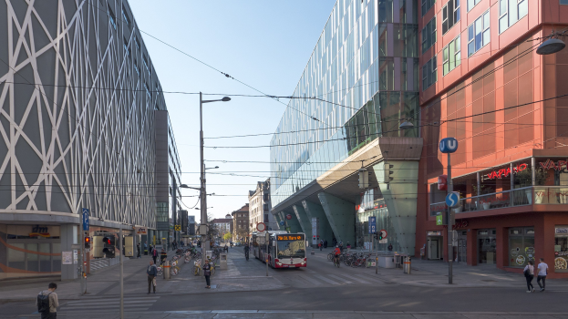 A city street with tall buildings, street poles, lights, electric cables, vehicles, pedestrians, bicycles, signs, and a clear blue sky.