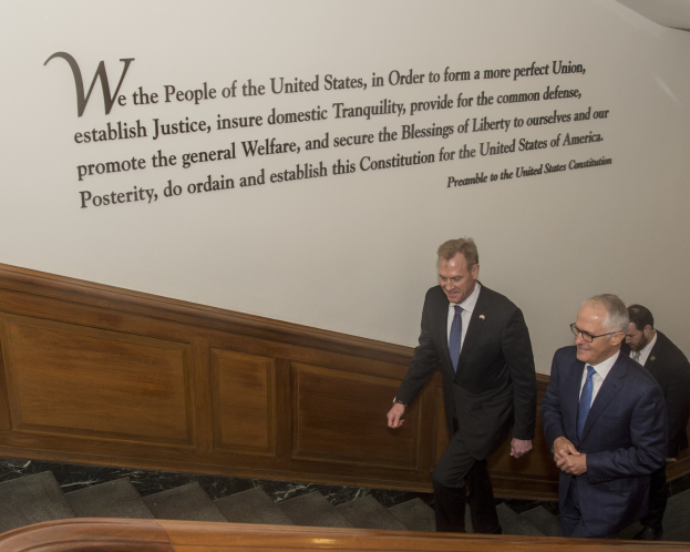 Two men in suits and ties walking down stairs, one wearing glasses, with a wall featuring text behind them.