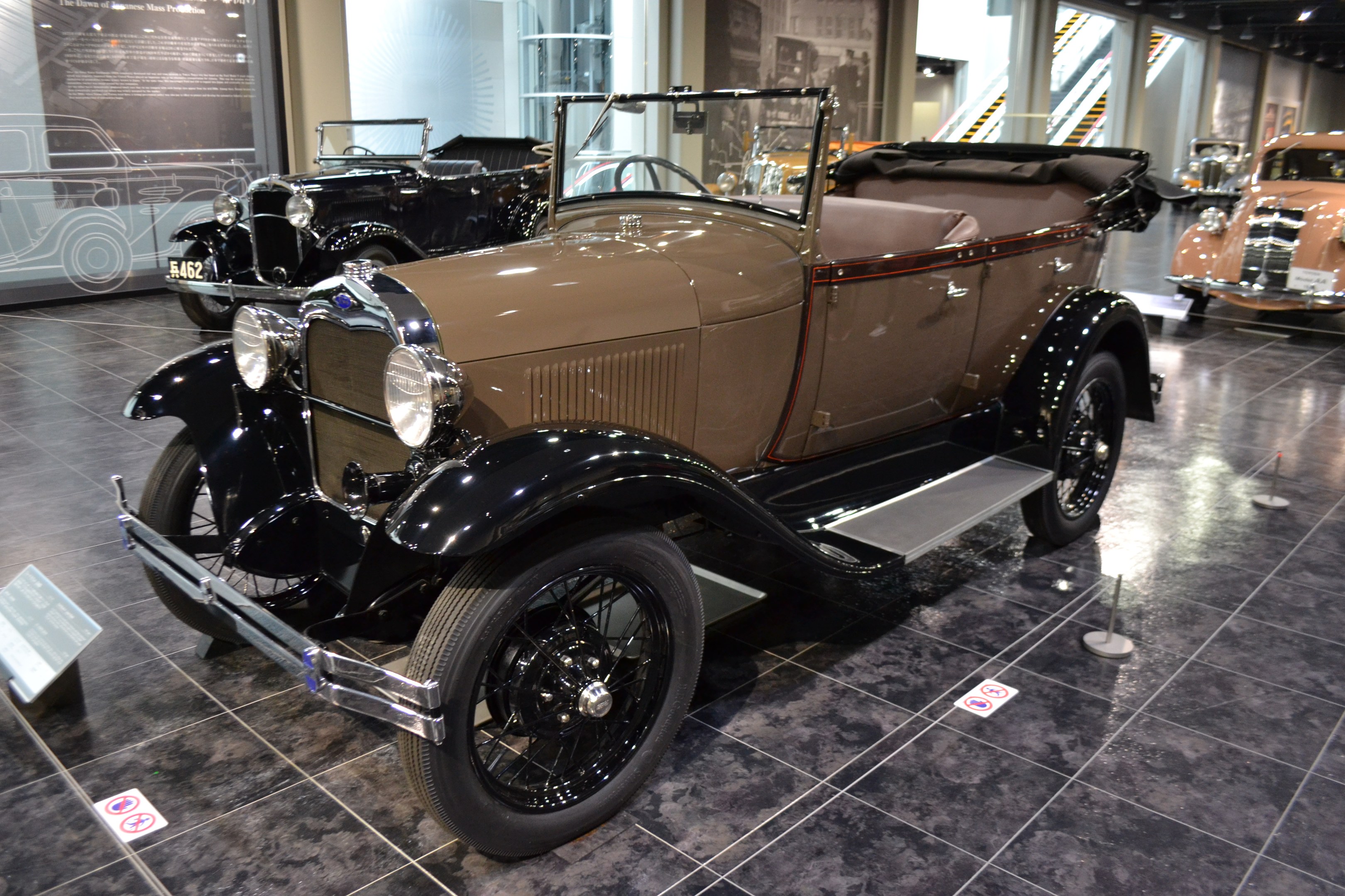 A vintage car displayed in a museum with informational boards, ribbon stands, glass walls, railing steps, and ceiling lights.