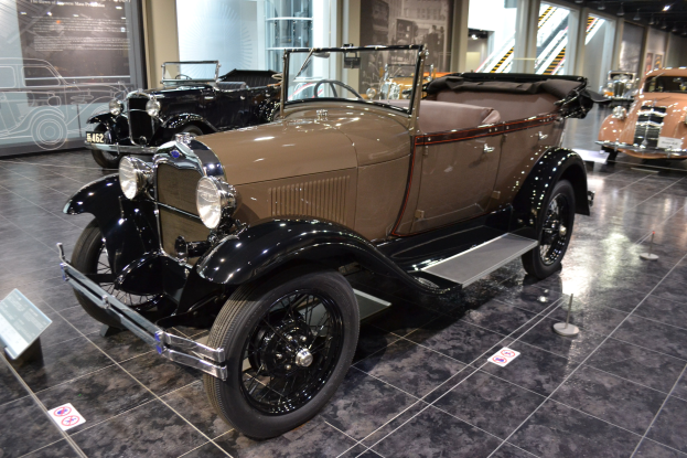 A vintage car displayed in a museum with informational boards, ribbon stands, glass walls, railing steps, and ceiling lights.
