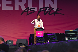 Ein Mann in einem weißen Hemd und braunen Hosen steht auf einer Bühne und hält ein Mikrofon in der Hand, mit einem pinken Banner mit der Aufschrift "Berlin as Fuck" dahinter und einer Menge davor.